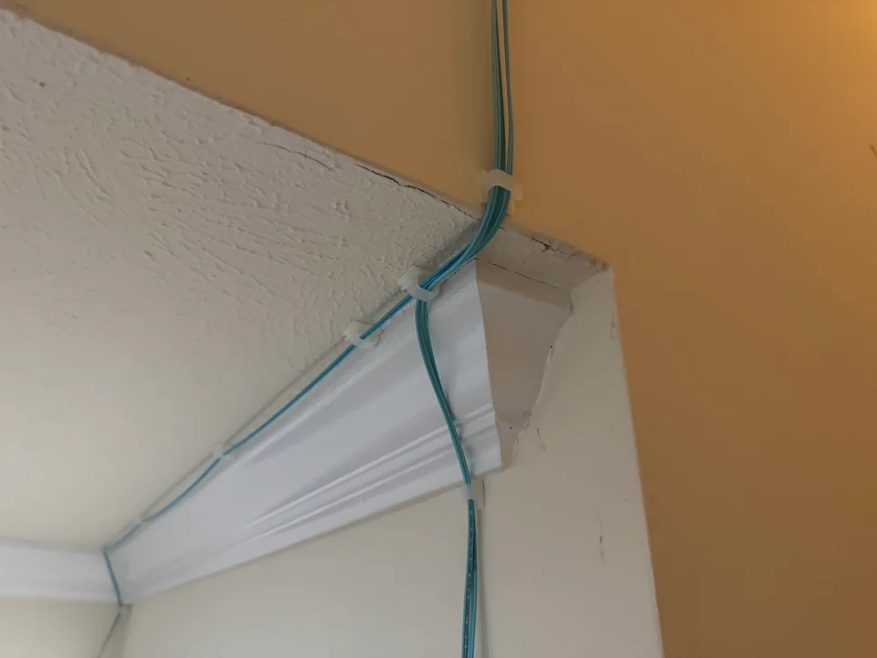 Aqua fiber optic cables running along white crown molding at a ceiling corner, secured with small white cable clips against orange-tan walls.